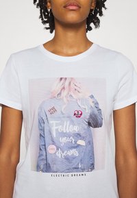 Camiseta blanca con una imagen impresa de una chaqueta de mezclilla con parches. El texto en la chaqueta dice "Sigue tus sueños" en una fuente divertida.
