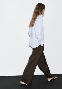Camicia azzurro chiaro con bottoni, vestibilità comoda; abbinata a pantaloni scuri marroni larghi e infradito nere, mentre cammina su una superficie nera.
