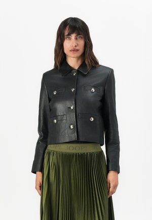 Frau trägt eine schwarze Lederjacke in cropped Schnitt mit silbernen Knöpfen und einen olivgrünen, hoch taillierten Faltenrock mit "JOOP!"-Markenkennung.