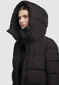 Chaqueta acolchada negra con una gran capucha plegable, tejido texturizado, costuras visibles y cierre de cremallera. Cuenta con cordones para ajuste.