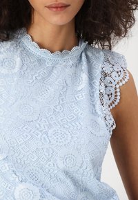 Ljusblå spetsblus med blommönster, vågig halsringning och spetsdetaljer på axlarna. Mjuk textur, semi-transparent tyg och intrikat design.