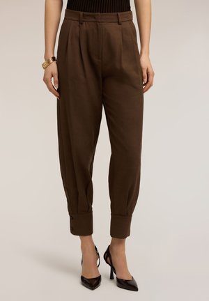 Kvinde iført brune bukser med høj talje og smalle ben med opbukket kant og knapdetaljer, kombineret med sorte højhælede sko med spids tå og gyldne armbånd.