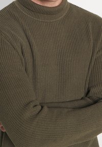 Olivgrön stickad tröja med ribbat mönster, hög krage och långa ärmar, med subtila variationer i stickningen.