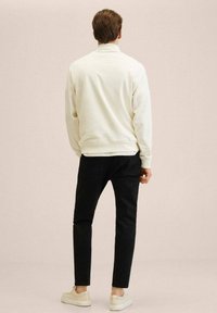 Sudadera blanca con dobladillo y puños acanalados, combinada con pantalones negros y zapatillas de color claro. Vista desde la parte posterior sobre un fondo neutro.