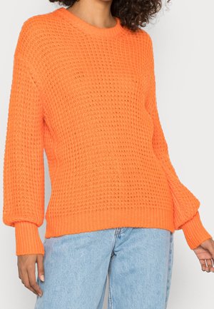 Pull en tricot de couleur orange vif, avec une coupe ample, des épaules tombantes, des poignets côtelés et un motif texturé sur l'ensemble.