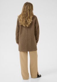 Cappotto lungo in misto lana marrone con taglio dritto, fessure laterali e cucitura posteriore pulita, abbinato a pantaloni beige a gamba larga e calzature nere.