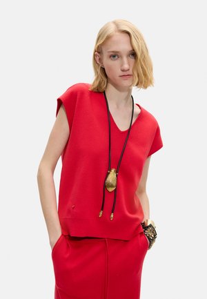 Blondynka w czerwonym bezrękawniku z dekoltem w kształcie litery V i spódnicy, nosząca długi czarny sznur z złotą zawieszką oraz bransoletkę z ćwiekami.