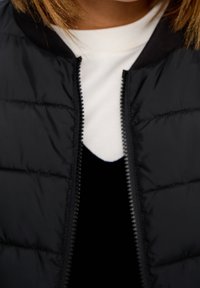 Donna con una camicia bianca indossata sopra un top nero senza maniche e una giacca a vento nera aperta, primo piano sul busto.