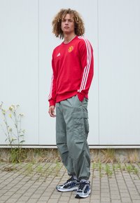 Rode sweatshirt met witte strepen, met een embleem op de borst. Olijfgroene cargo pants met zijzakken. Zwarte en blauwe sport schoenen.