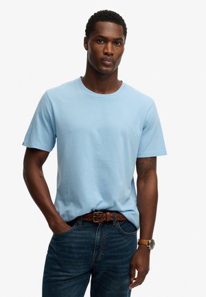 Mann trägt hellblaues kurzärmliges T-Shirt, dunkelblaue Jeans mit braunem geflochtenem Gürtel, Armbanduhr, steht vor weißem Hintergrund.