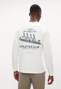 Bílé mikina s dlouhým rukávem s grafickým designem kreslených postaviček pádlujících na kajaku, s nápisy "SKUTEČNÝ STOCKHOLM" a "KLUB ATLETŮ."