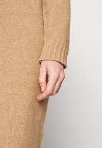 Close-up van een hand die naast een persoon ligt die een camel-kleurige gebreide trui met geribde manchetten draagt.