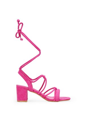 Sandaña de tacón cuadrado de suede rosa con tiras delgadas y ataduras en el tobillo, que presenta una punta cuadrada y una textura suave.