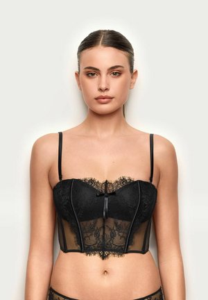 Mujer con el cabello peinado hacia atrás, vestida con un bralette negro de encaje con tirantes delgados, de pie contra un fondo claro y liso.