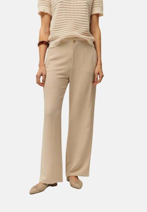 Mujer que lleva pantalones anchos beige, una camiseta de punto crema de manga corta, bailarinas tejidas beige y una pulsera marrón en su muñeca izquierda.