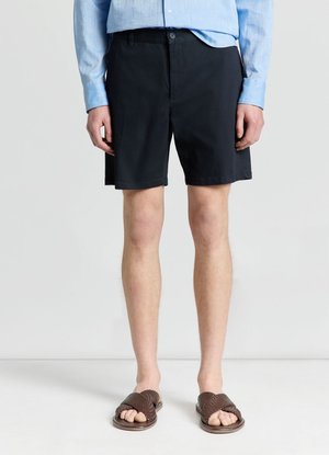 Uomo che indossa una camicia azzurro chiaro a maniche lunghe, pantaloncini neri fino al ginocchio e sandali intrecciati marroni, in piedi su un pavimento bianco davanti a uno sfondo bianco.
