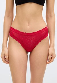 Γυναίκα  που φοράει κόκκινο δαντελωτό μπικίνι εσώρουχο χαμηλής μέσης και μαύρο κοντό τοπ, στέκεται με τα χέρια χαλαρά στα πλευρά σε απλό φόντο.