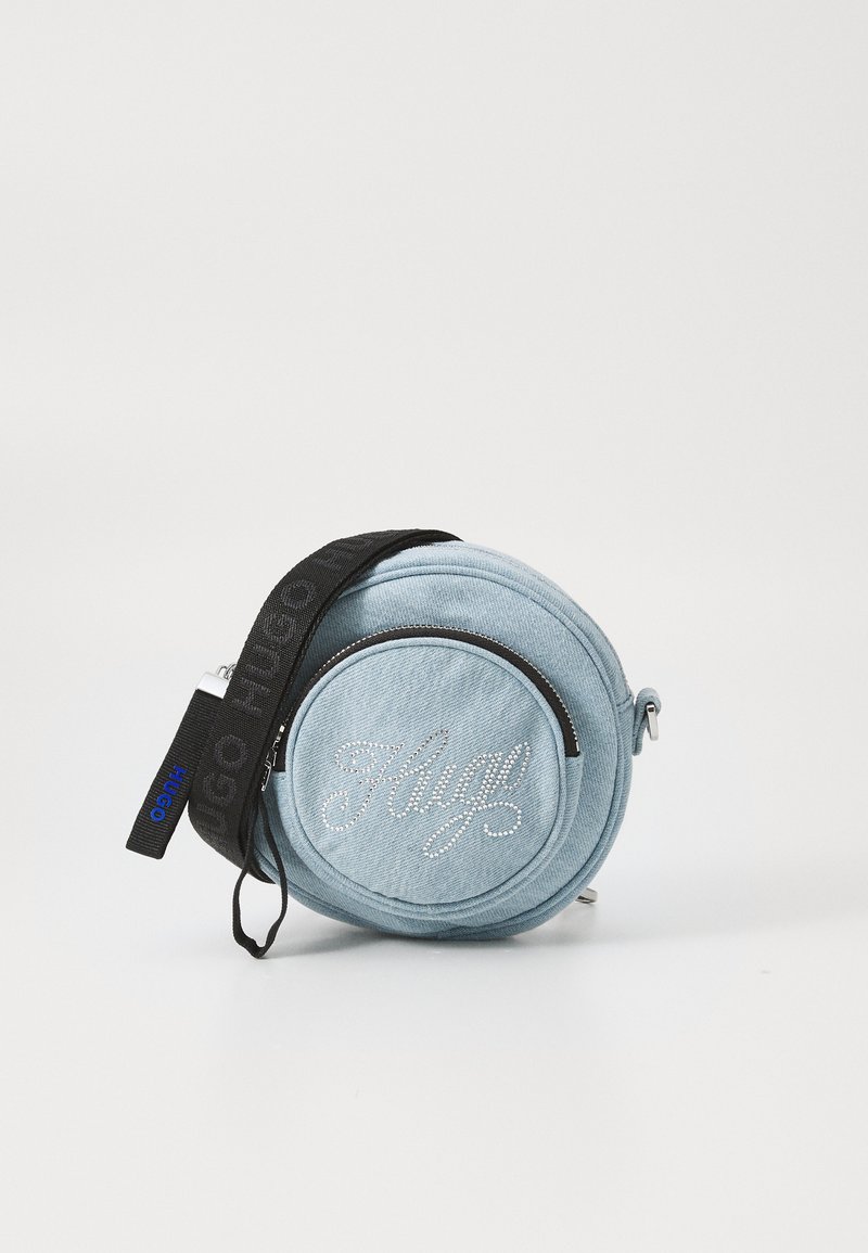 Sac rond en denim bleu clair avec une bandoulière noire et une fermeture éclair argentée, présentant un design en relief "Hugo" en strass sur le devant.