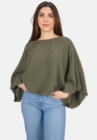 Olivengrünes, übergroßes Top aus strukturiertem Stoff, mit weiten Ärmeln und dekorativem Spitzenbesatz am Saum, kombiniert mit hellblauen Jeans.