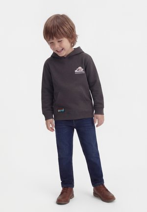 Young boy smiling, wearing dark gray Quiksilver hoodie, blue jeans, and brown boots, standing against a plain light background.