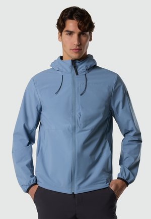 Man wearing a light blue zip-up hooded jacket with pockets, standing with hands in pockets, facing forward against a plain background.