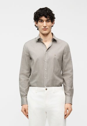 Homme portant une chemise à motifs beige boutonnée rentrée dans un pantalon blanc, se tenant devant un fond uni clair.