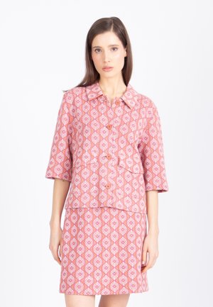 Žena oblečená v ružovej vzorovanej bundovej blúzke s rukávmi do lakťov a zodpovedajúcej sukni po kolená na bielom pozadí.