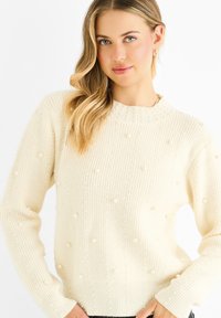 Cream-colored knitted sweater featuring a ribbed texture with textured details and pearl-like accents distributed throughout. Round neckline.