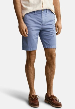 Mann trägt hellblaue knielange Shorts, beige kurzärmeliges Hemd und braune geschnürte Lederschuhe, steht vor weißem Hintergrund.