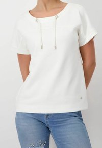 Valkoinen lyhythihainen paita, jossa on leveä kaula-aukko, nyöritys-detail ja hopeanväriset yksityiskohdat. Sileä kangas, rento istuvuus. Yhdistetty sinisiin farkkuihin.