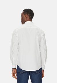 Camicia bianca con bottoni e maniche lunghe, realizzata in tessuto liscio. Presenta un colletto classico e un orlo curvo, abbinata a jeans blu scuro.