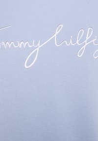 Světle modrá látka s bílým vyšitým nápisem "Tommy Hilfiger." Materiál vypadá měkký a má hladkou texturu.