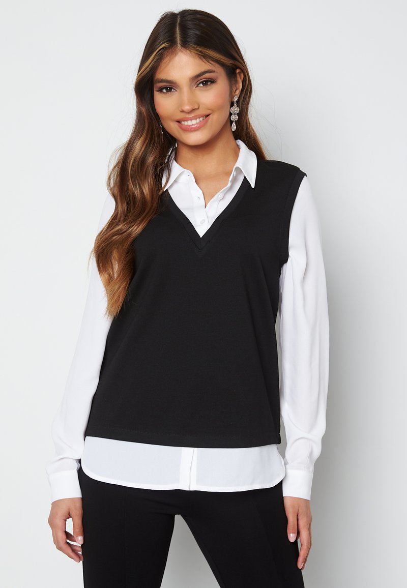 Maglione senza maniche nero con scollatura a V, indossato sopra una camicia bianca con bottoni. Presenta un tessuto liscio e un design aderente.