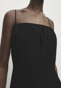 Schwarzes ärmelloses Kleid mit gerafftem Ausschnitt und feinen Trägern. Glatte Textur, A-Linien-Silhouette und minimalistische Designdetails.