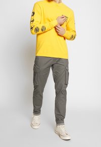 Gelbes Langarmhemd mit schwarzen Grafiken an den Ärmeln, kombiniert mit grauen Cargo-Hosen und beigen Sneakern mit weißen Sohlen.