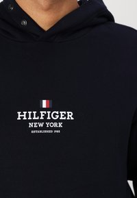 Hoodie de color azul marino oscuro de algodón con cordón, que presenta un logo blanco "HILFIGER NEW YORK" y un pequeño detalle de bandera en rojo, blanco y azul.