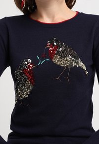 Maglione blu navy con decorazioni di uccelli di paillettes; due uccelli si fronteggiano, uno tiene una bacca rossa con foglie verdi nel becco.