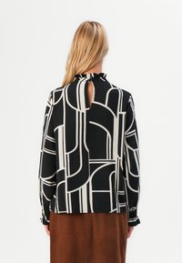 Donna con lunghi capelli biondi che indossa una blusa nera e beige con un motivo geometrico e una piccola apertura a chiave sul retro, abbinata a una gonna marrone.