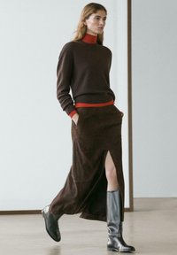 Frau, die drinnen geht und einen braunen Pullover über einem roten Rollkragen, einen braunen Rock mit Schlitz und schwarze kniehohe Stiefel trägt.