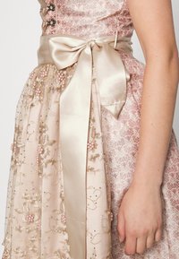 Robe à motif floral en tissu rose pâle avec broderies dorées et accents de perles. Ruban en satin noué en un nœud à la taille. Texture délicate.