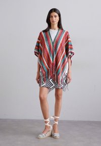 Poncho tejido colorido con patrón de zigzag en rojos y teal, con bordes con flecos; llevado sobre un vestido a rayas blancas y negras y espadrilles.