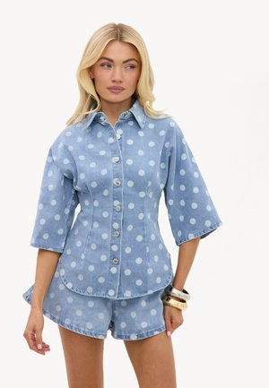 Femme blonde portant une chemise en denim bleu clair et un short à pois blancs, debout avec des bracelets à son poignet sur fond blanc.