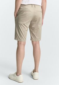 Person, der beigen knielangen Shorts und weiße Sneaker trägt, steht mit den Händen in den Taschen vor einem schlichten weißen Hintergrund.
