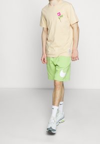 Beżowa koszulka z bawełny z różowym logo i zielonym wzorem liścia, w komplecie z jasnymi zielonymi szortami z dużym białym logo Nike. Białe sneakersy.