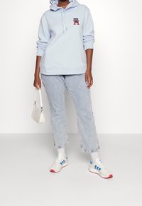 Sudadera azul claro con un logo, combinada con jeans rectos de lavado claro y zapatillas blancas con acentos azules y rojos. Llevando una pequeña bolsa blanca.