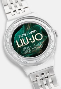 Sølv metalurur med en cirkulær urskive, sort og grøn digital skærm, omkranset af ædelstene, med et metalarmbånd og krystal detaljer.