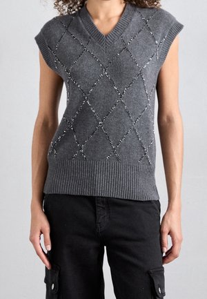 Femme portant un gilet gris en tricot sans manches avec un motif en losange et un pantalon cargo noir, debout devant un fond uni.
