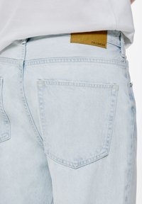 Jeans din denim de culoare albastră deschisă, cu o textură fină, având o etichetă din piele de culoare bej pe talie și două buzunare la spate.