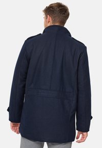 Navy wool blend coat with a high collar, shoulder epaulettes, and back vent, featuring a smooth texture and tailored shape.