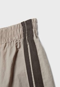 Pantalon à taille élastique beige avec double rayure verticale marron foncé sur le côté, gros plan sur la texture du tissu et les coutures.
